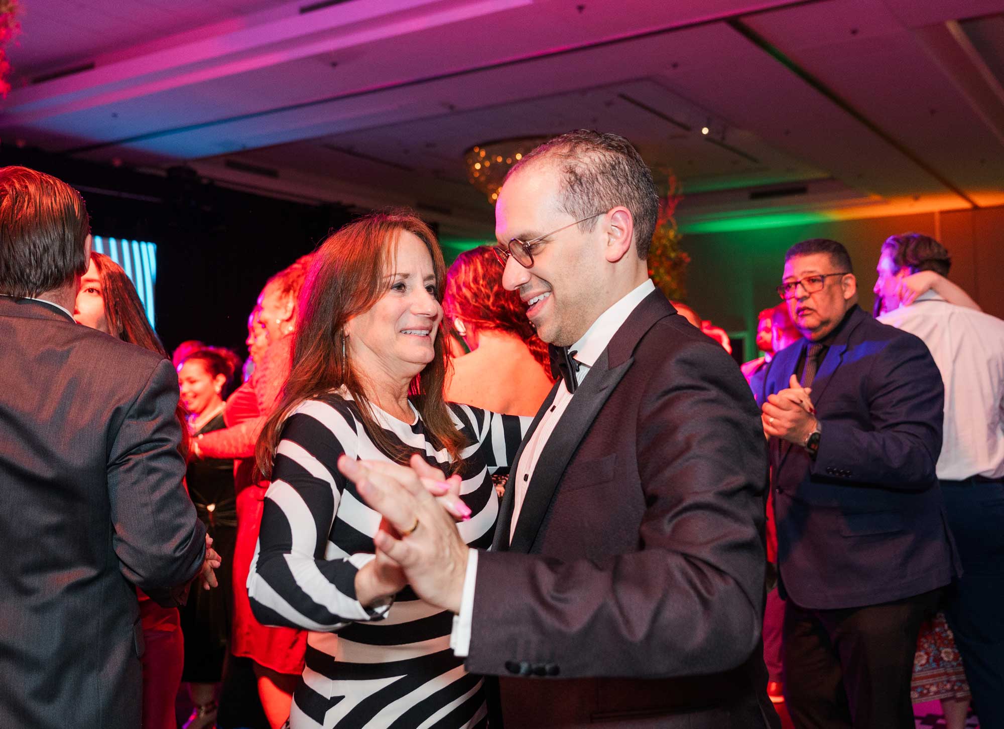 A man in a suit and a woman in a black-and-white striped dress dance together at a crowded indoor event, with other people dancing in the background.