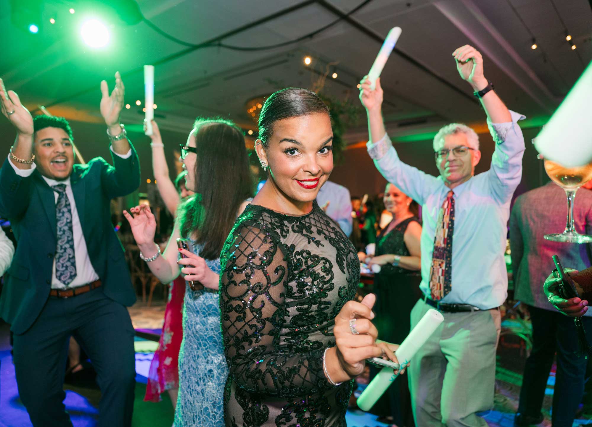 A woman in a black lace dress stands at the center of a lively indoor party, surrounded by people dancing and holding glow sticks under colorful lights.