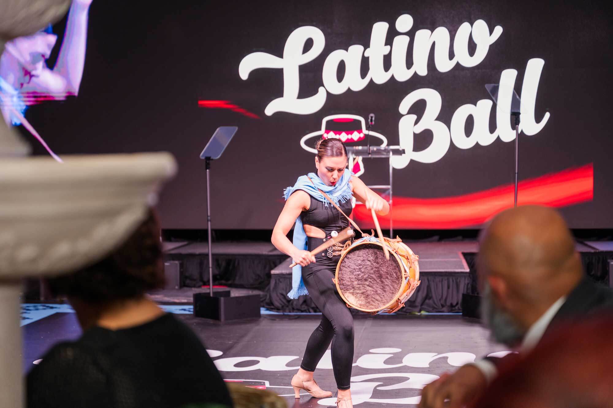 A performer in black attire plays a drum on stage at the Latino Ball, with the event name displayed in large letters behind her.