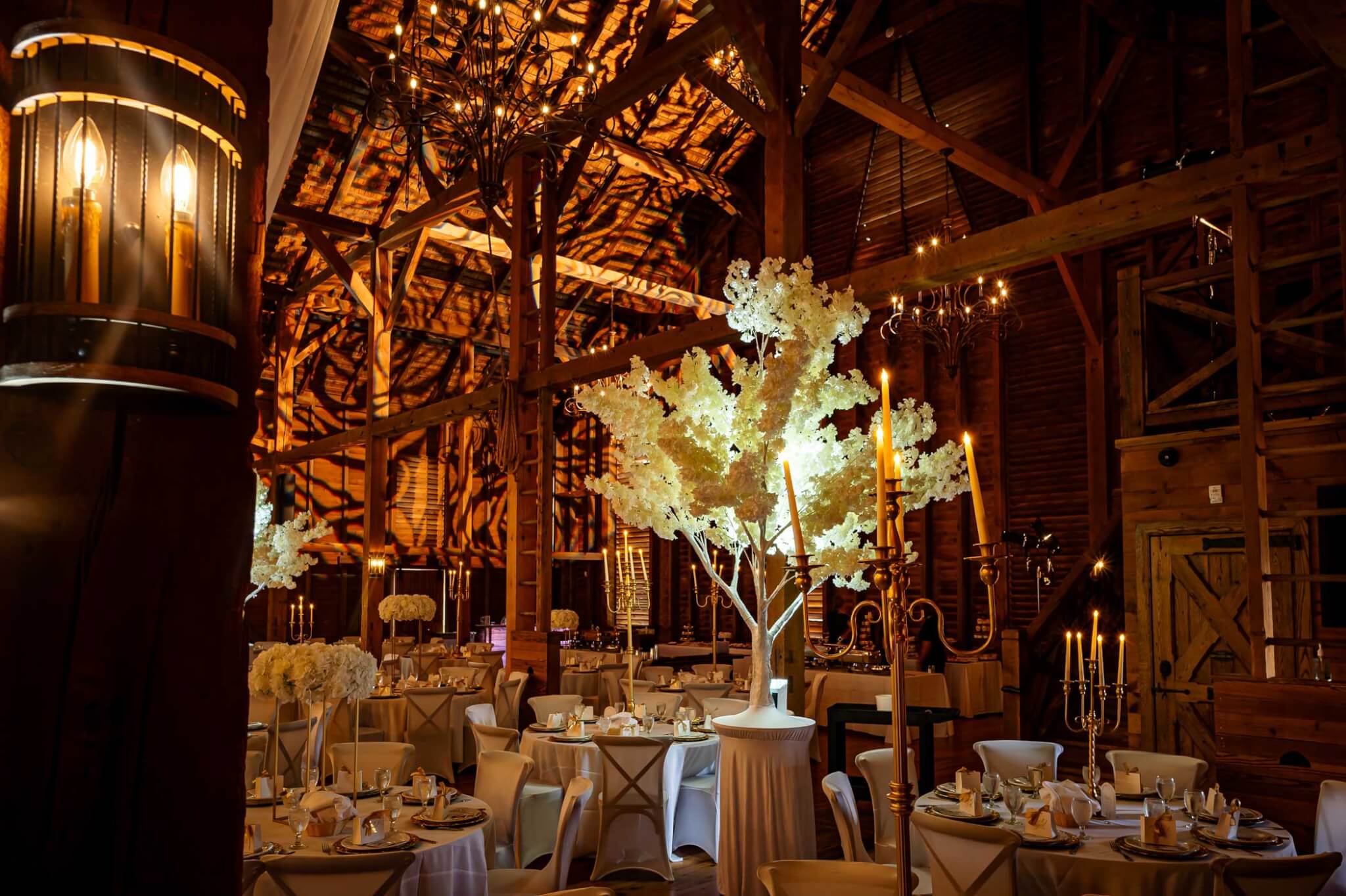 A decorated barn venue with round tables, white floral centerpieces, glowing candles, and warm ambient lighting highlighting wood beams and chandeliers.