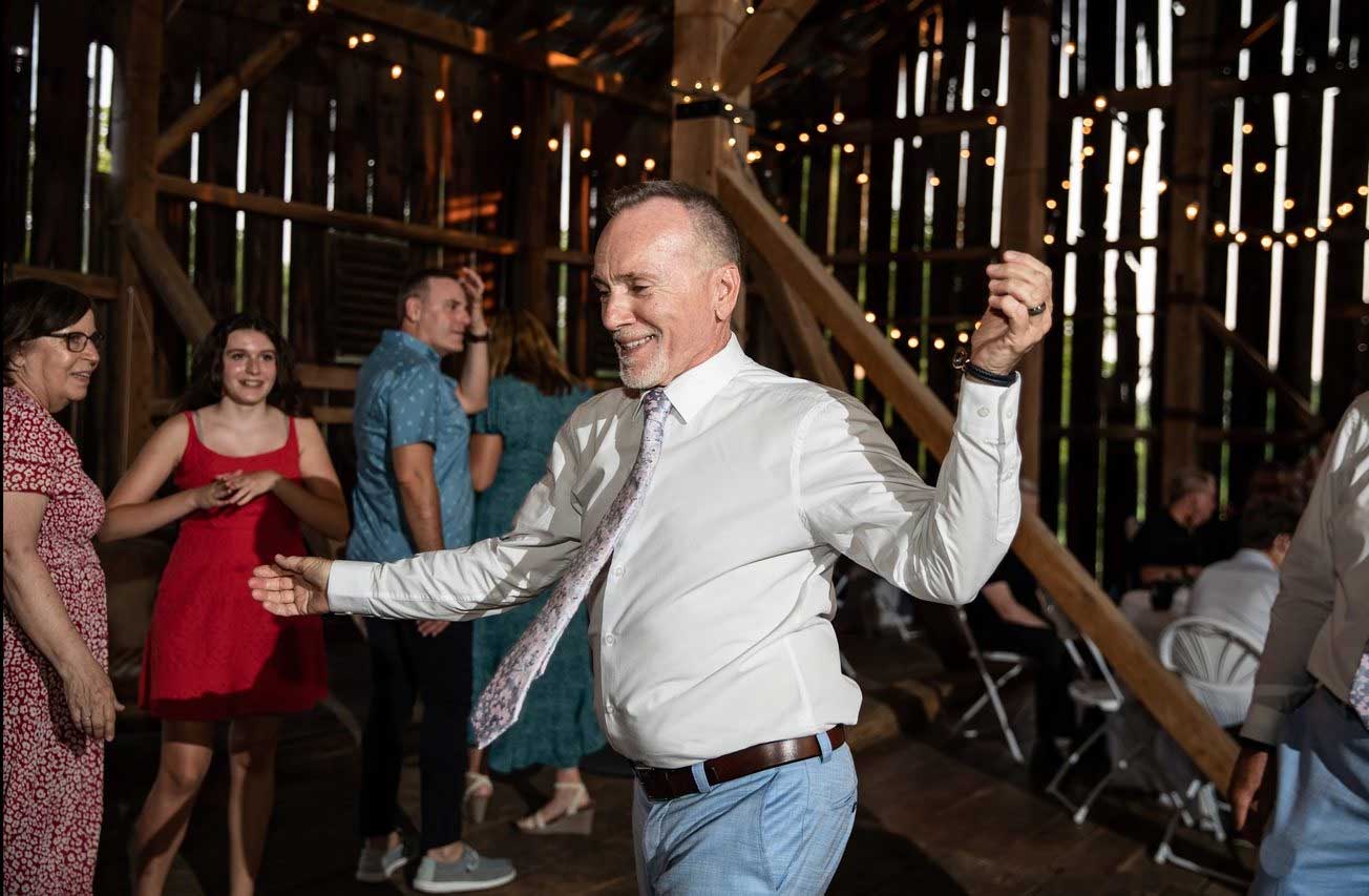 man-dancing-at-barn-party-event A man in a white shirt and tie dances in a rustic barn with string lights, while other people socialize and watch in the background.