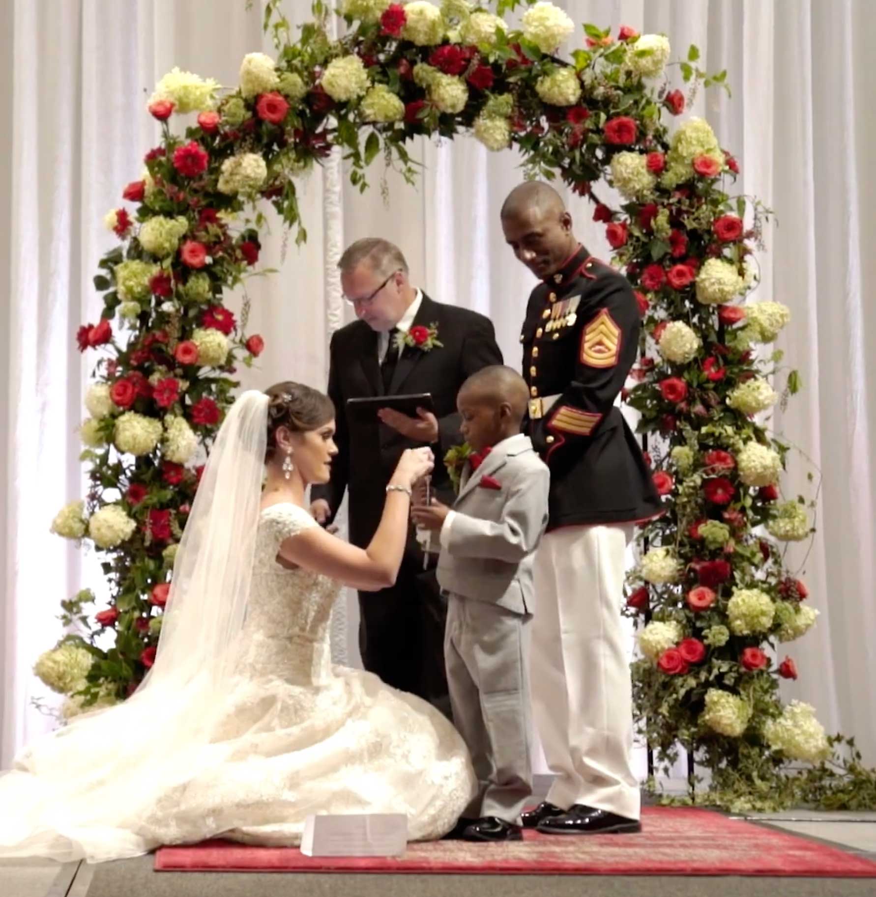 military-wedding-altar-with-child A bride kneels in front of a young boy in a suit, with a groom in military uniform and an officiant standing under a floral arch.