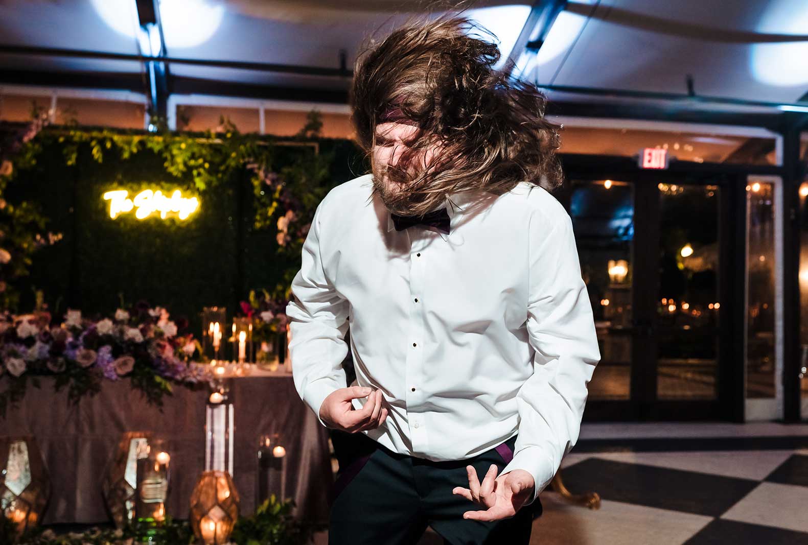 party-man-dancing-air-guitar-hair A man in a white dress shirt with long hair flips his head and pretends to play air guitar at an indoor event with floral decorations and candles in the background.
