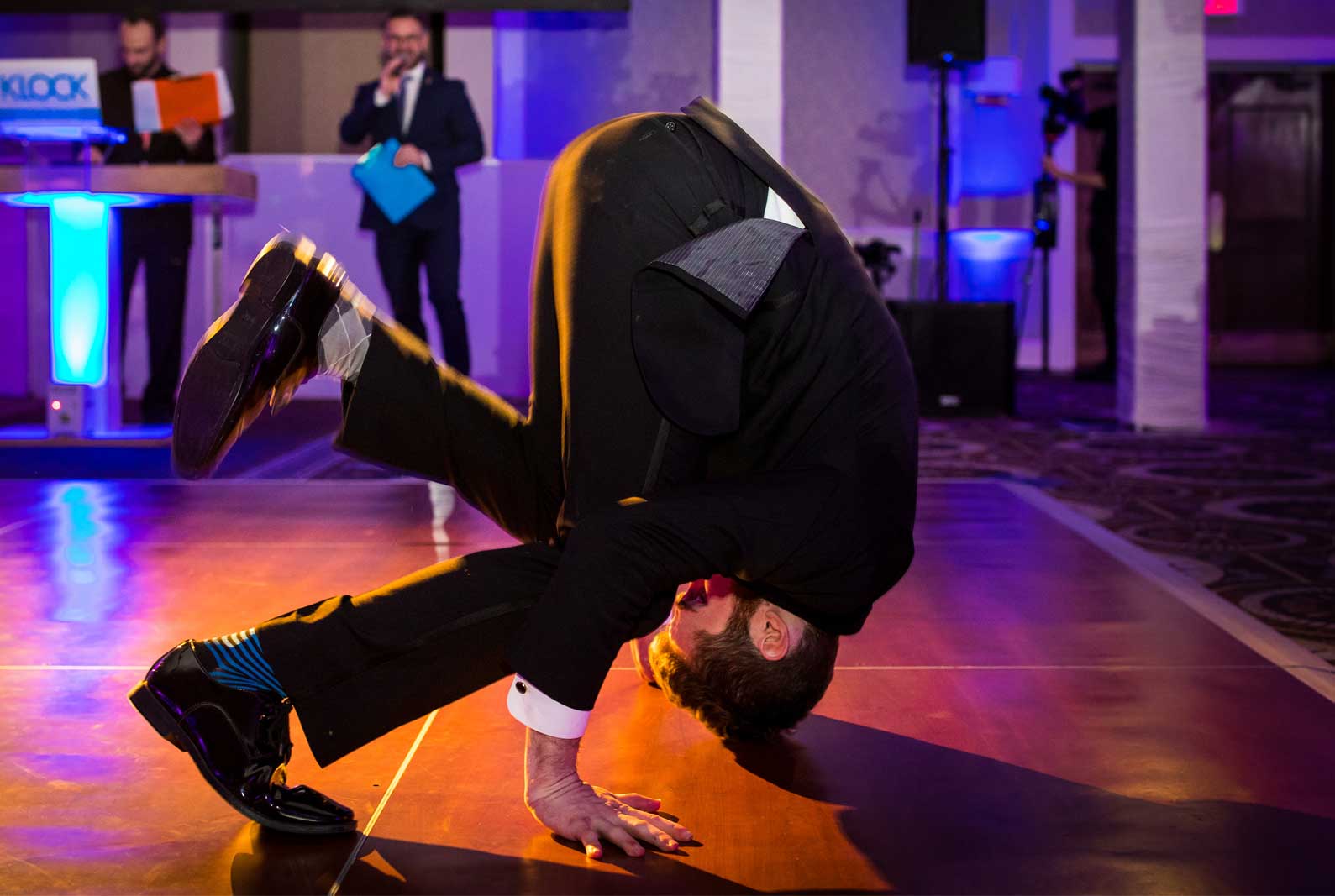party-man-dancing-on-head-dance-floor A man in a suit balances on his hands and head on a dance floor at an indoor event, while others watch in the background.