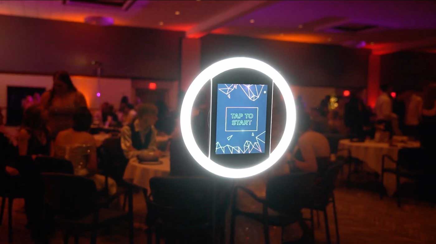 photo-booth A digital photo booth with a circular LED light and a screen reading "Tap to Start" stands in a dimly lit event room with people seated at tables in the background.