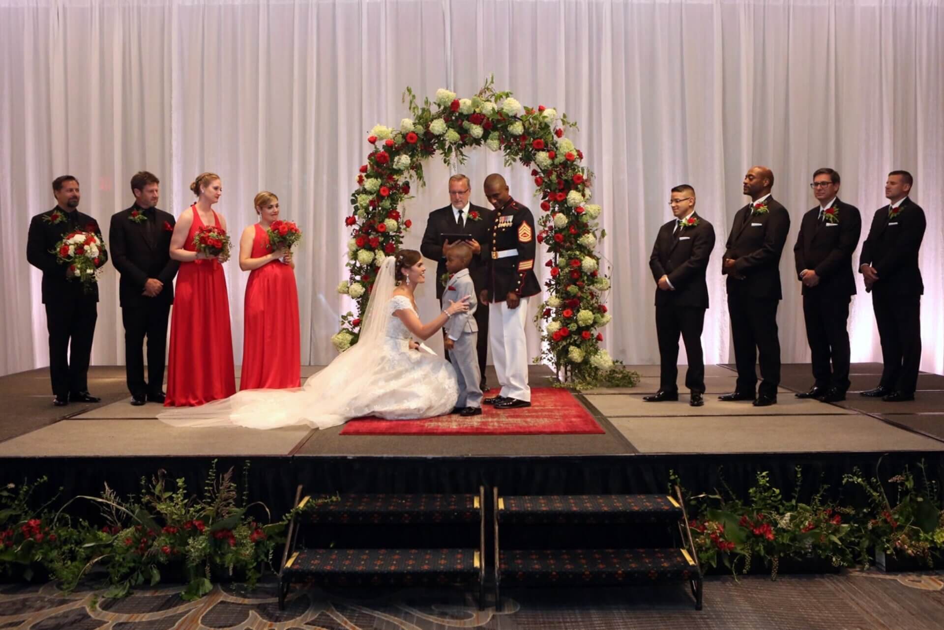 Military-wedding-altar-with-child A bride and groom kneel at the altar during a wedding ceremony, surrounded by bridesmaids in red dresses and groomsmen in black suits on a decorated stage.
