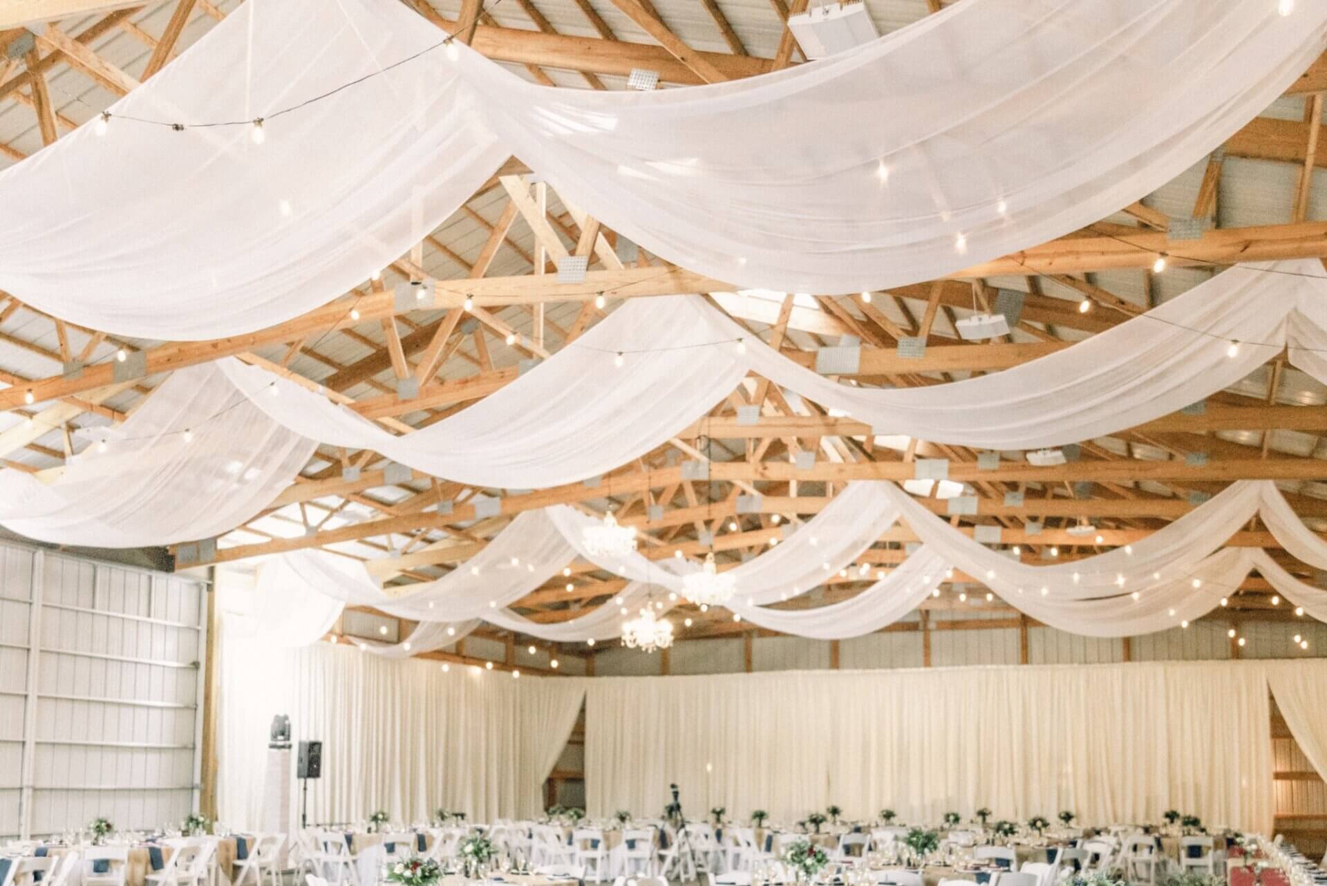 A spacious event hall with wooden beams, draped white fabric, hanging string lights, and rows of round tables with white chairs set for a gathering.