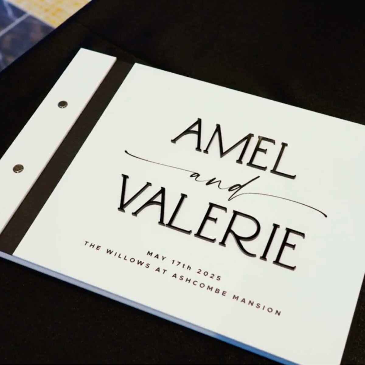 A white guestbook with “Amel and Valerie” in large text, and “May 11th, 2025, The Willows at Ashcombe Mansion” written below, lies on a black surface.