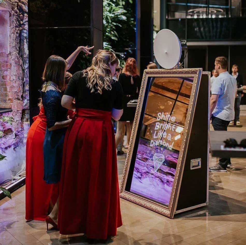 Three women in formal dresses stand in front of a large interactive mirror displaying the text "Shine Bright Like a Diamond" at an indoor event.