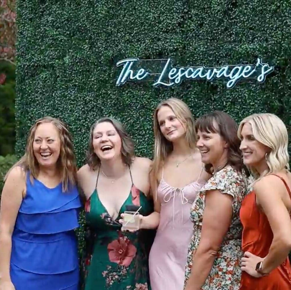 Five women posing and smiling in front of a green hedge wall with a neon sign that reads "The Lescavage's.