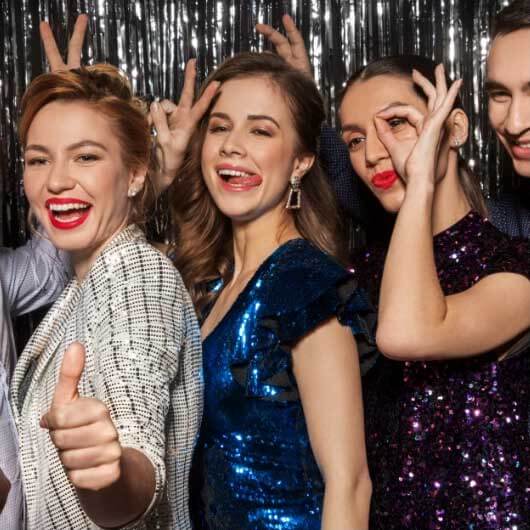 Five people in party attire stand in front of a silver tinsel backdrop, smiling and making playful hand gestures toward the camera.