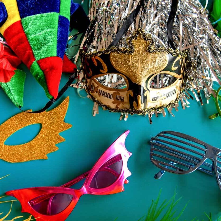 A variety of colorful party accessories, including masks, novelty glasses, a jester hat, leis, metallic fringe, and feathers, arranged on a turquoise surface.