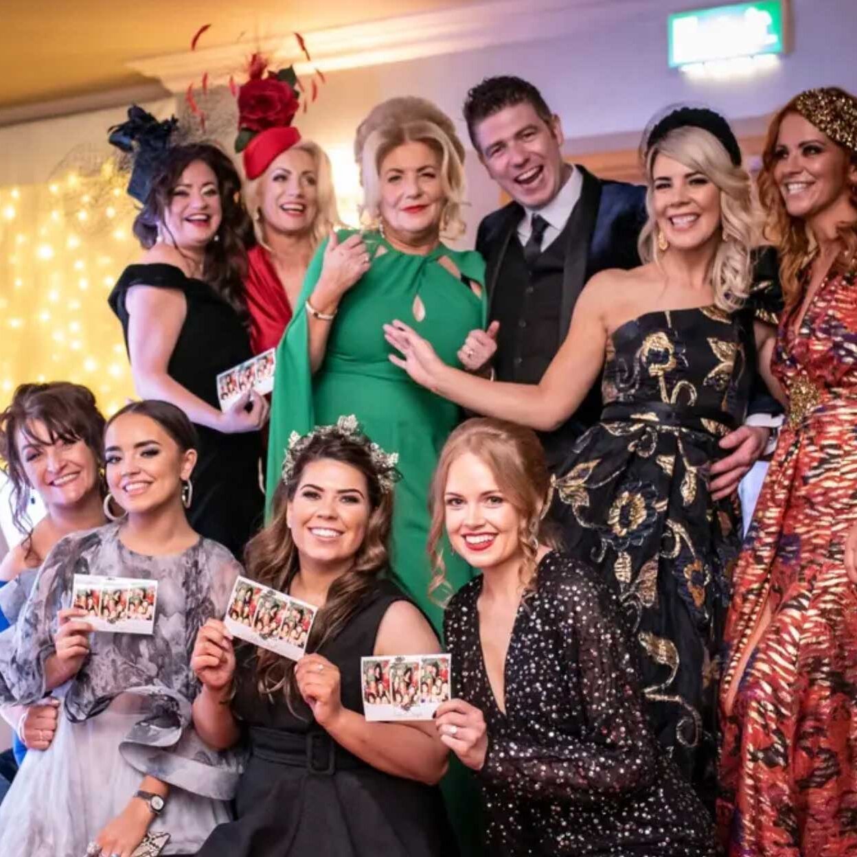 A group of well-dressed people, some holding cards, pose and smile for a photo at an indoor event with decorative lights in the background.