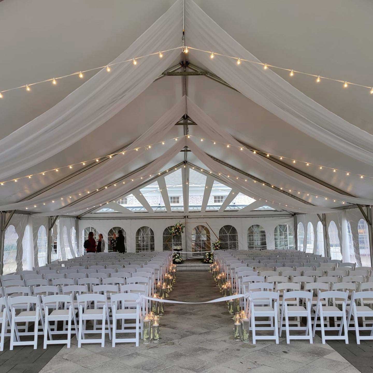 A wedding ceremony setup under a white tent with rows of white chairs, hanging lights, draped fabric, and floral arrangements along the aisle.