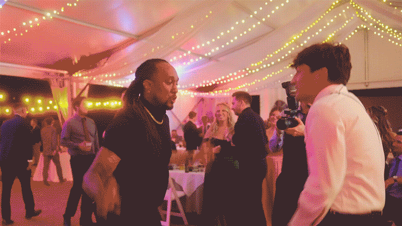 video-for-klock-mobile Two men are talking and dancing at a lively indoor event under string lights, with other people mingling in the background.