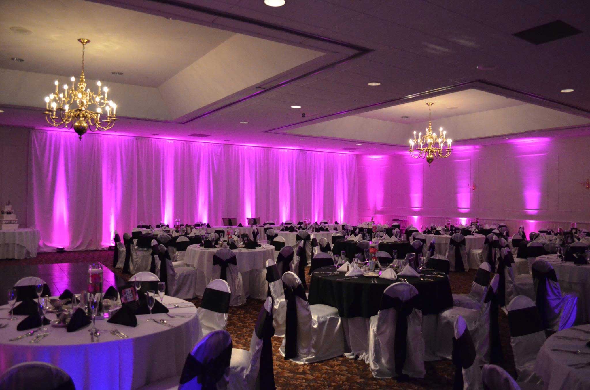 A banquet hall set for an event, with round tables and chairs covered in white linens, purple uplighting on the walls, and chandeliers hanging from the ceiling.