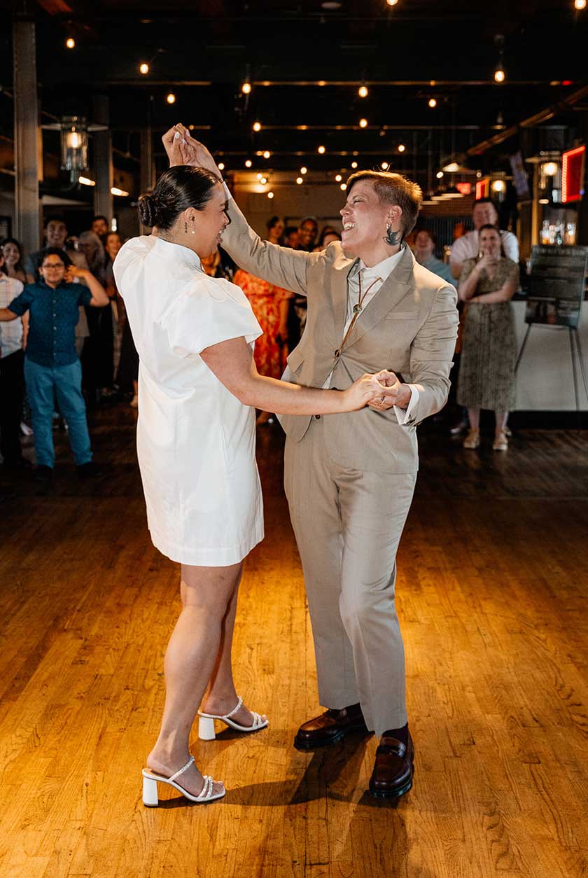 wedding-dance-lgbtq-The-Abbey-Bar Two people smile and dance together in a warmly lit room, surrounded by onlookers. One wears a white dress, the other a tan suit.