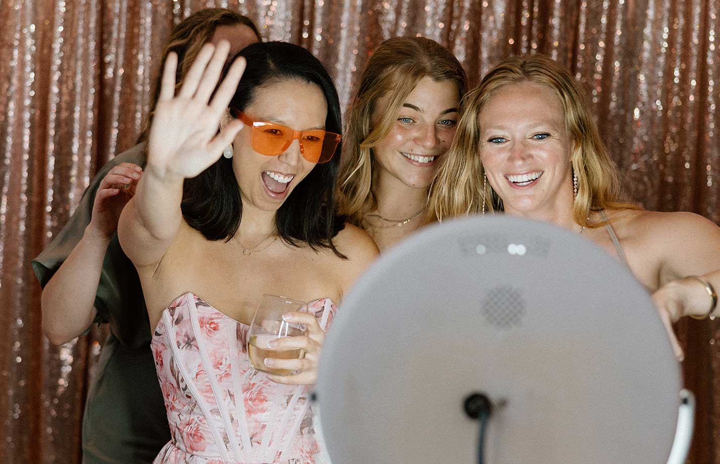 women-at-photo-booth Four women pose and smile in front of a photo booth with a sequined backdrop; one woman is waving, holding a drink, and wearing orange sunglasses.