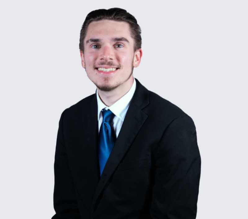 Chris_Berry A young man in a black suit, white shirt, and blue tie sits and smiles at the camera against a plain light background.