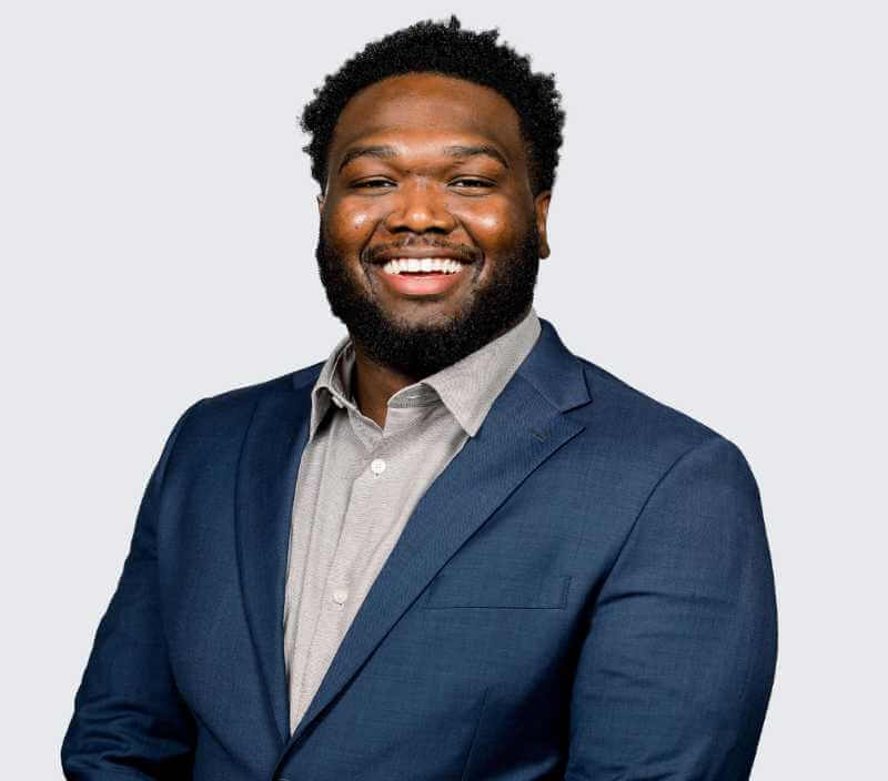 phips_Jules A man in a dark blue suit and light gray shirt smiles while standing against a plain light background.