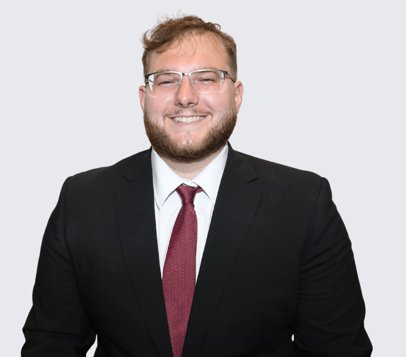 Andrew-Bongiorno A man with short hair, glasses, and a beard is smiling while wearing a black suit, white shirt, and red tie, standing against a plain light background.