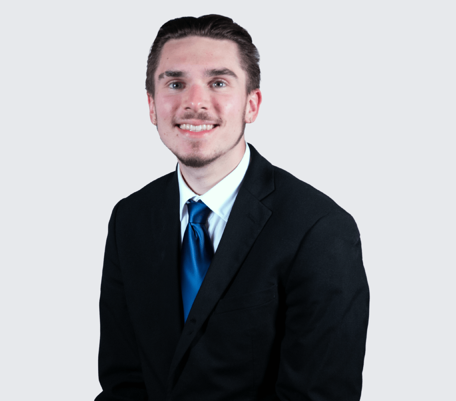A young man in a black suit, white shirt, and blue tie is sitting and smiling at the camera against a plain light background.