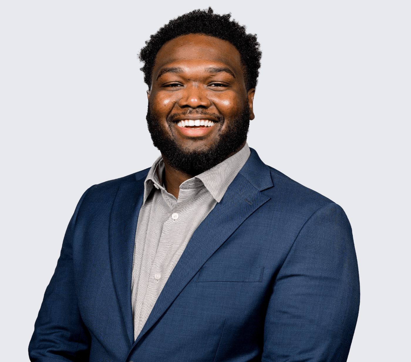 A man with a beard and short curly hair smiles, wearing a blue suit jacket and light-colored shirt, posed against a plain light gray background.