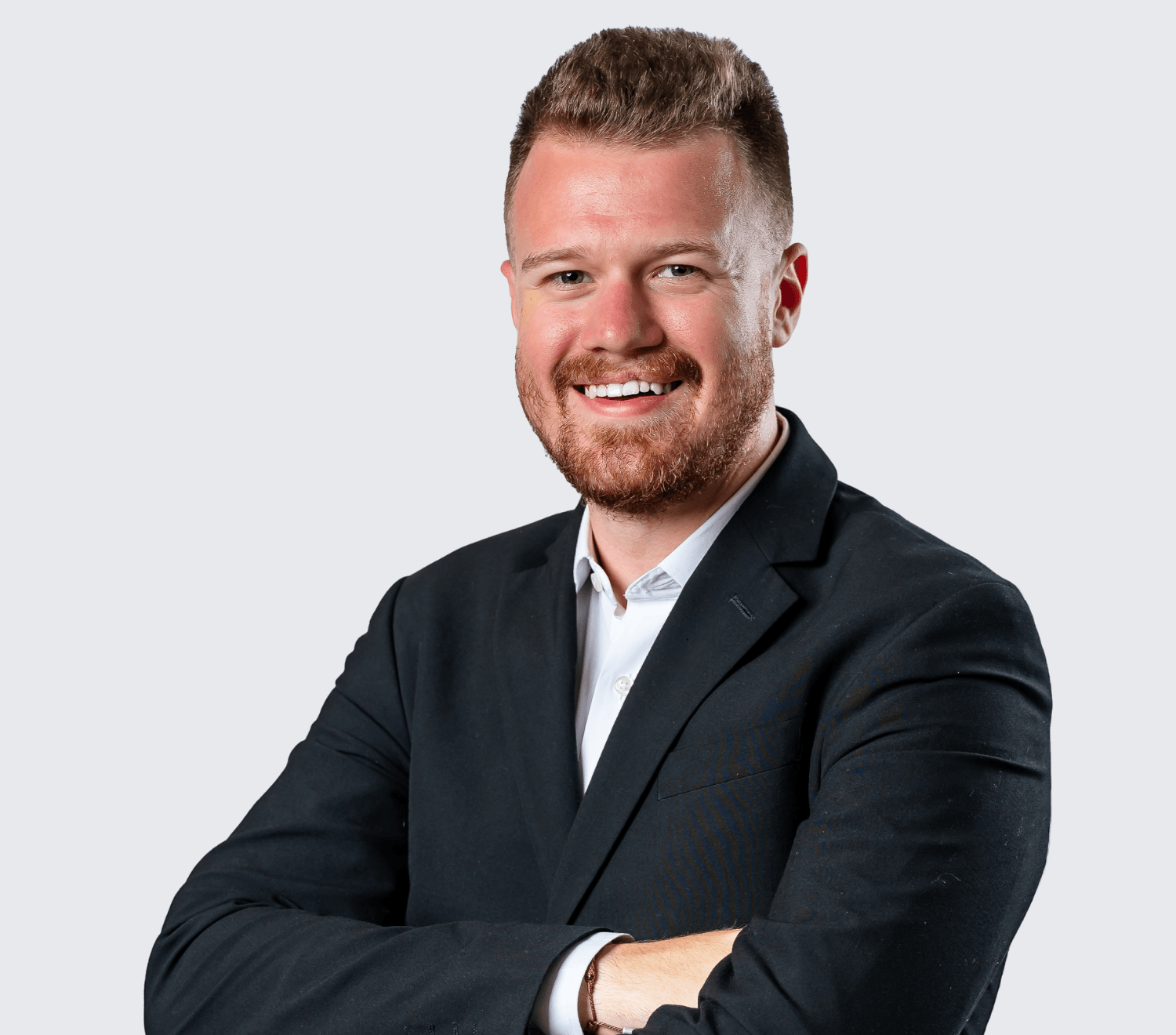 Man with short hair and beard in a black suit jacket and white shirt, smiling with arms crossed against a plain light background.
