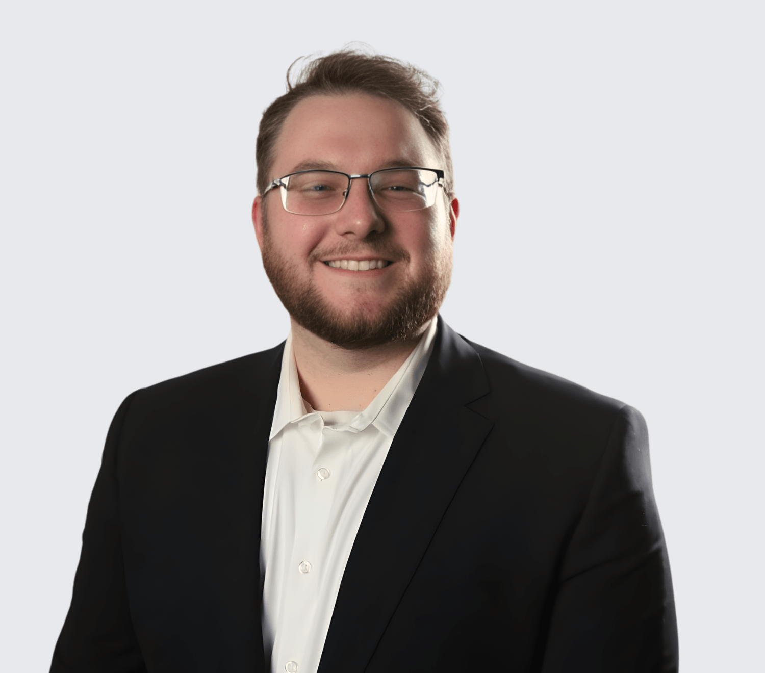 A man with glasses, a beard, and short hair is smiling while wearing a white shirt and a dark blazer against a plain light background.