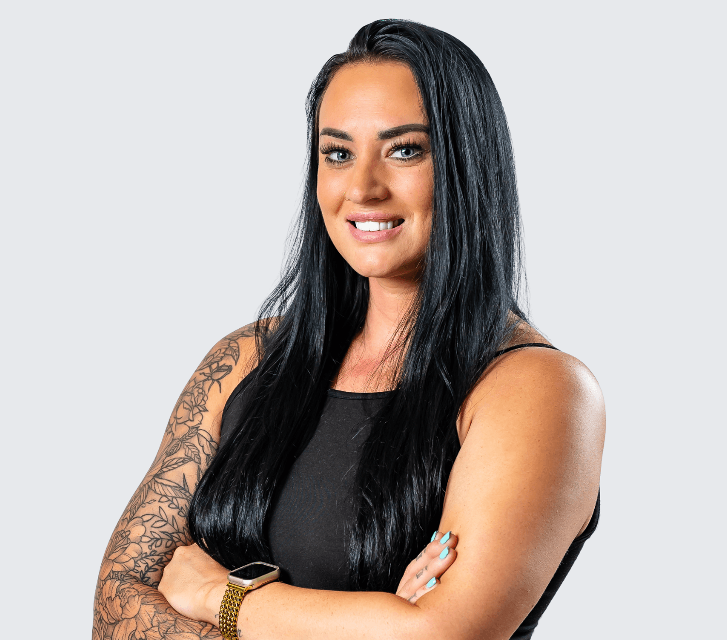 Woman with long black hair, wearing a sleeveless black top and a smartwatch, stands with arms crossed and smiles at the camera against a plain light gray background.