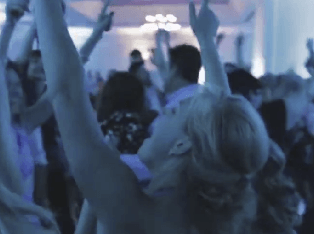 A crowd of people stands close together indoors, raising their arms and hands in the air under blue lighting.