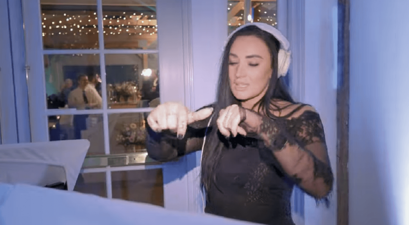 DJ Micheh in a black lace dress and headphones stands indoors, gesturing with her hands in front of DJ equipment. String lights and people are visible through the window behind her.