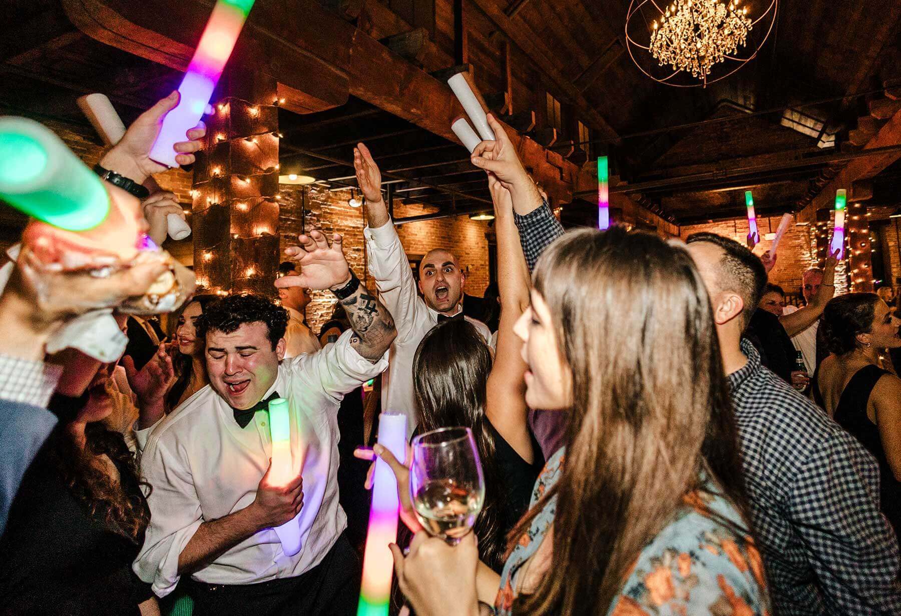 Wedding guests dancing with glow sticks under string lights and a chandelier at a Klock Entertainment reception