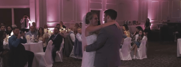 A bride and groom share their first dance at a wedding reception, surrounded by seated guests in a decorated hall with pink lighting at colonial country club