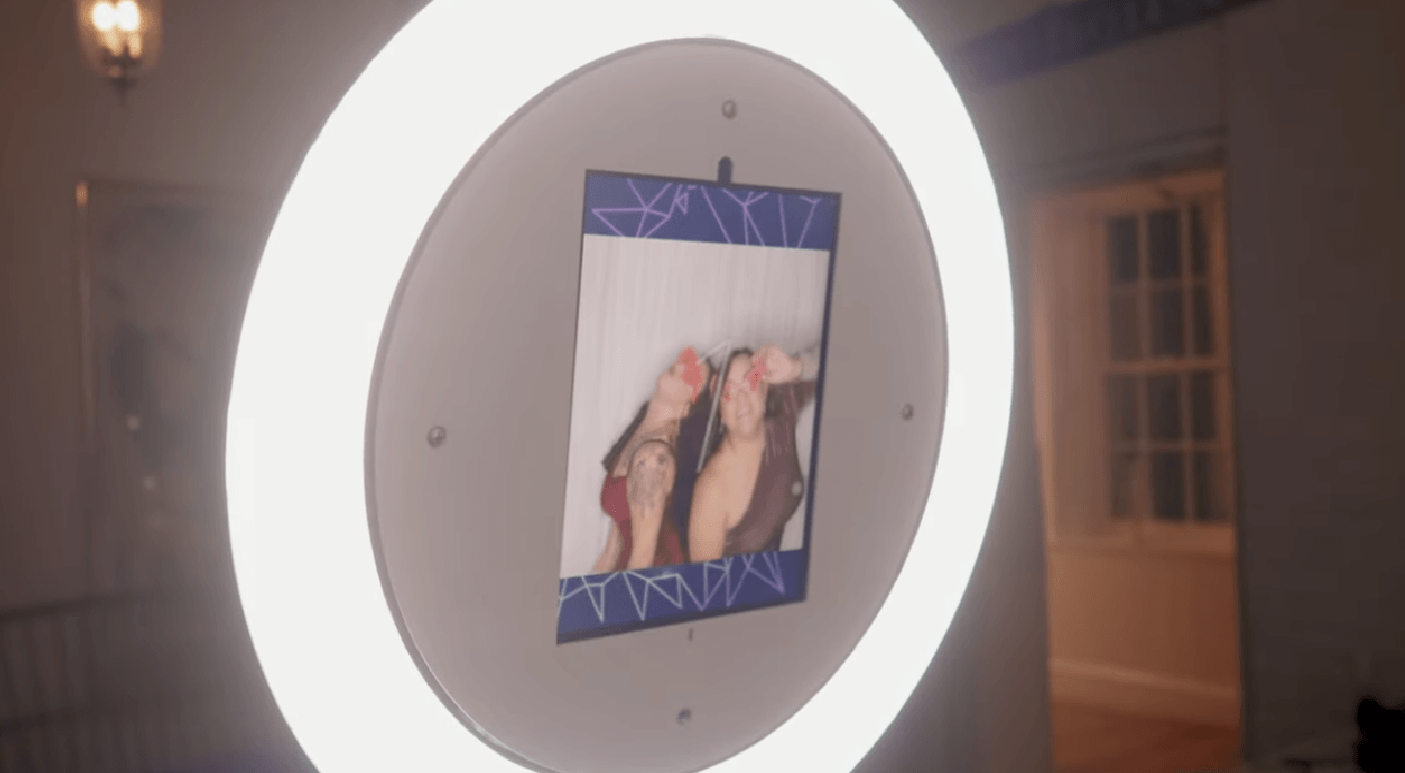 A circular ring photo booth frames a printed photo of two people making silly faces, displayed on a photo booth setup indoors.