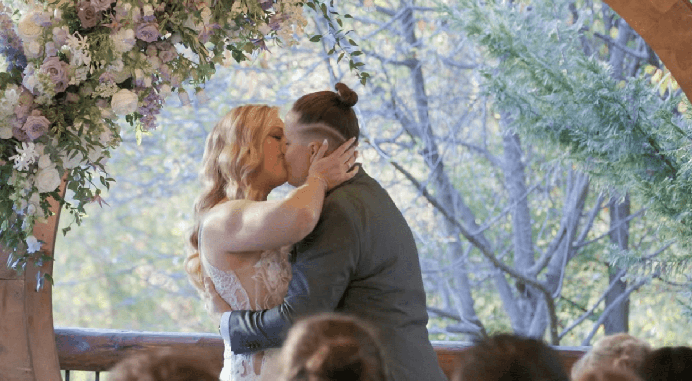 Two people kiss under a flower arch at an outdoor wedding ceremony sound offering, surrounded by trees as guests watch.