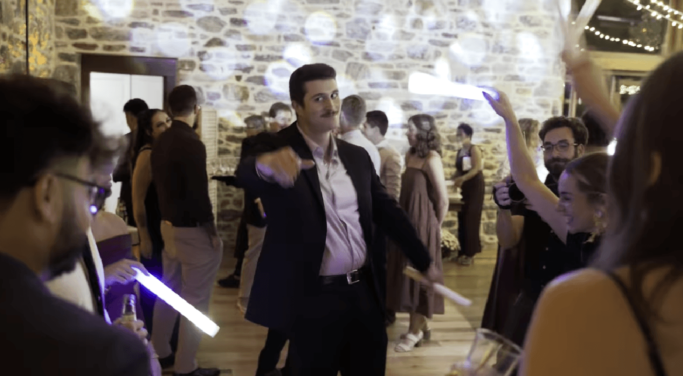 wedding dancing bluestone estate A group of people in formal attire dance indoors with glow sticks, surrounded by string lights and a stone wall at bluestone estate.