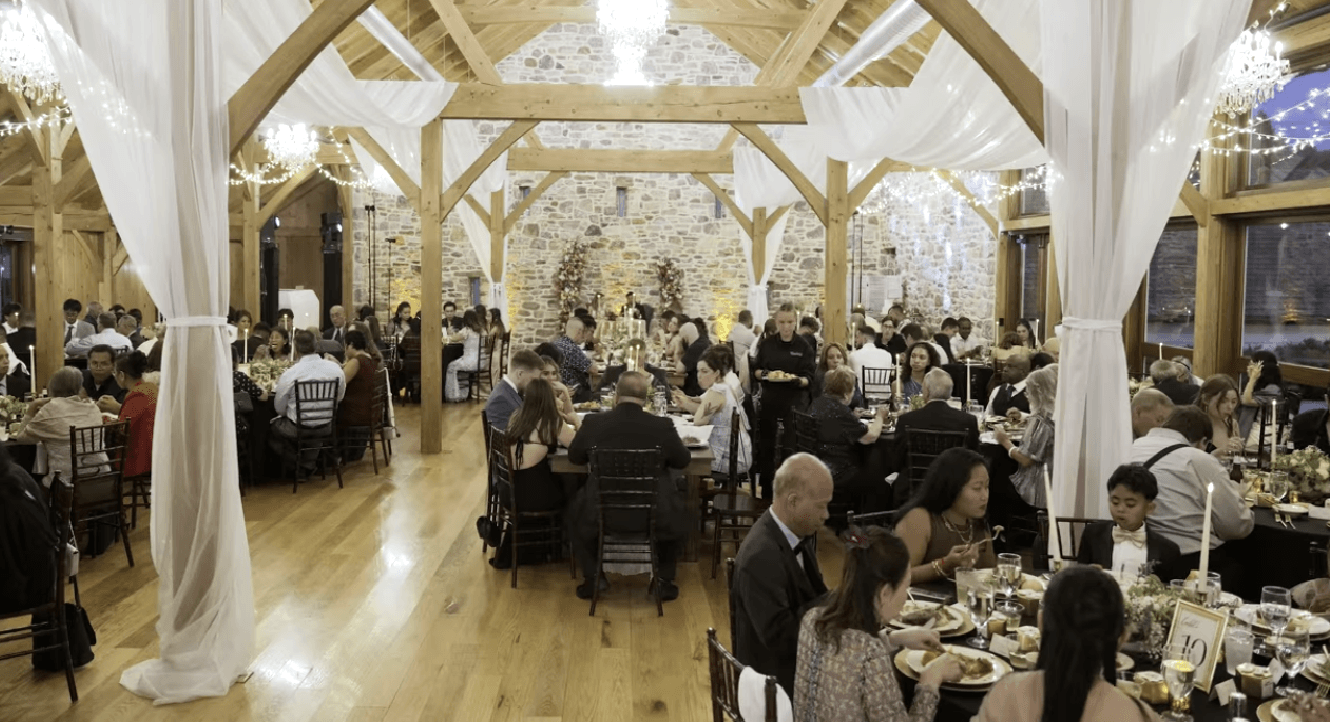 wedding dinner bluestone estate A formal dinner event is held in a rustic hall with wooden beams, stone walls, round tables, and string lights at bluestone estate.