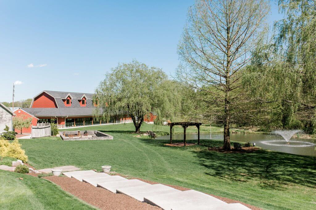 A large red barn-style event building next to a grassy lawn, a pond with a fountain, and a wooden gazebo at Historic Acres of Hershey. 