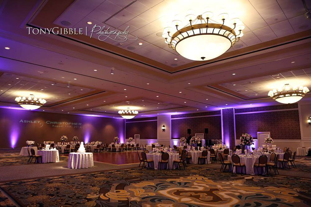 A large ballroom set for a wedding reception with round tables, floral centerpieces, and purple uplighting.