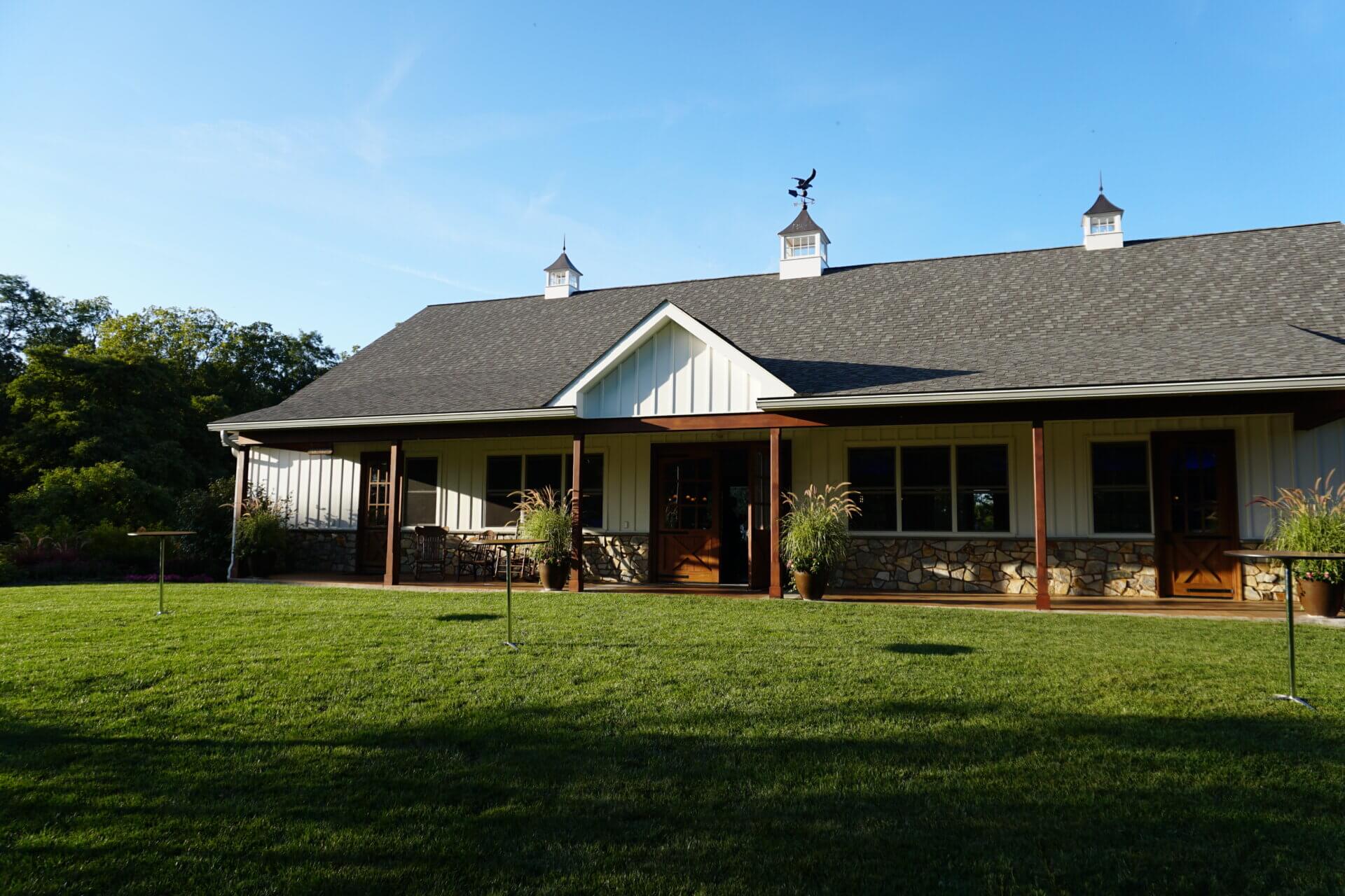 A single-story white barn-style building with a stone base, a dark shingled roof with cupolas, and a covered porch facing a large grass lawn at Drumore Estate