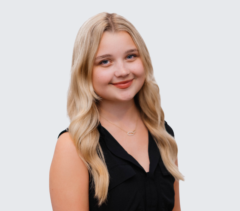 Portrait of a young woman with blonde hair wearing a black sleeveless top against a light background.