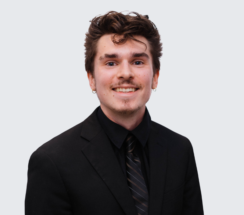 Chris-26 Headshot of a smiling young man wearing a black suit, black shirt, and a dark striped tie.