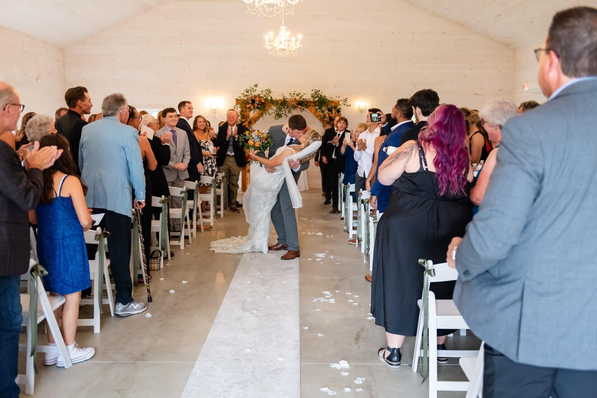 A bride and groom kiss while dipping on the aisle of a wedding ceremony as guests look on and applaud at Farm 1840