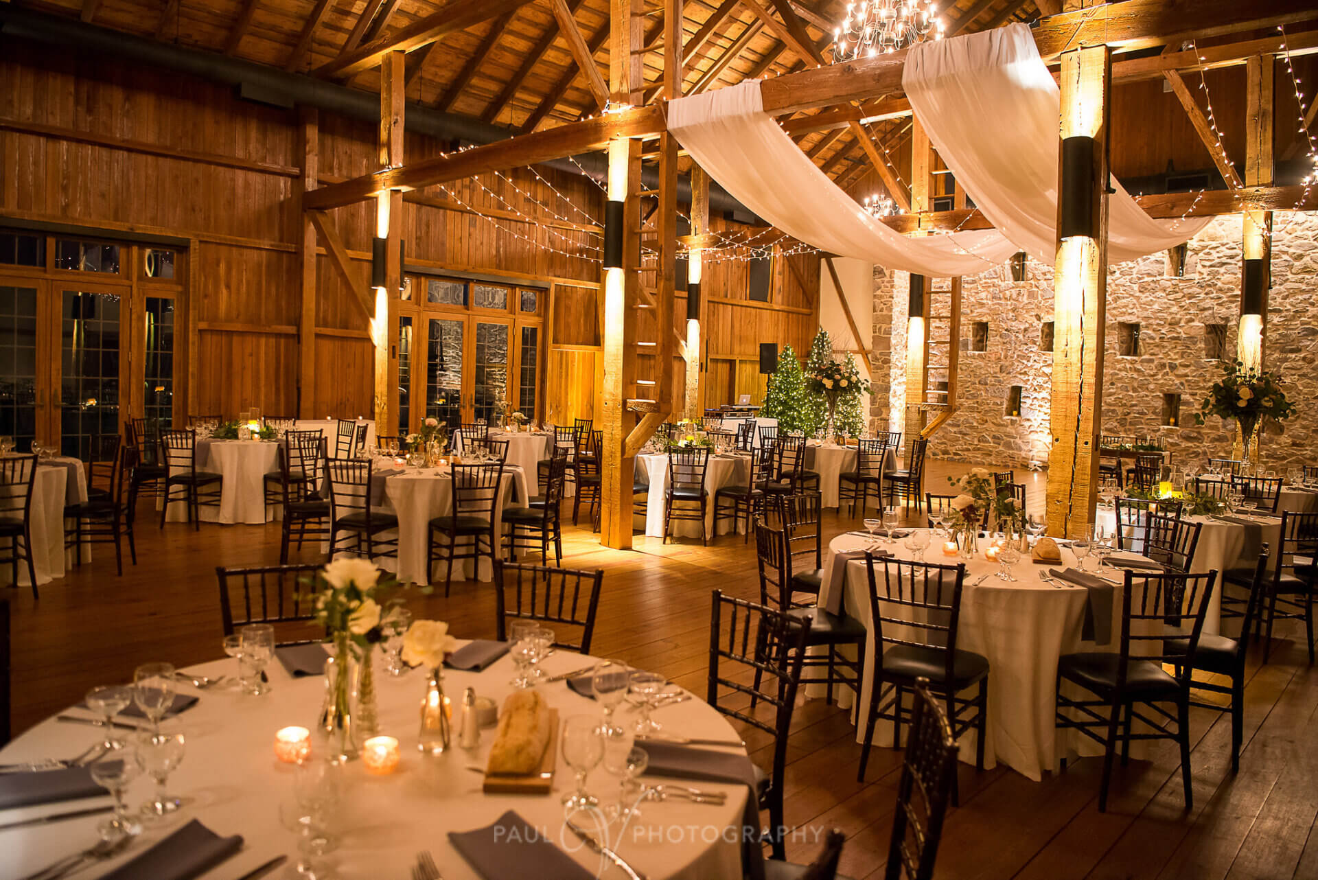 A rustic barn wedding reception with round tables set for dinner, draped white fabric, and string lights hanging from the wooden ceiling beams at Harvest View Barn. 