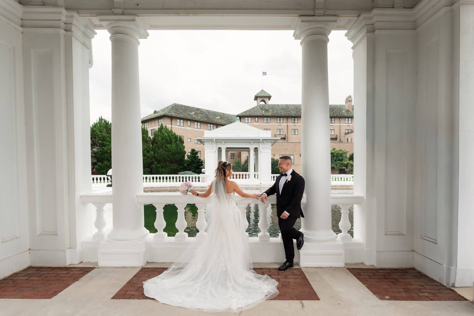 A bride and groom pose together while leaning on the railing of a white pavilion overlooking a pond and Hotel Hershey.