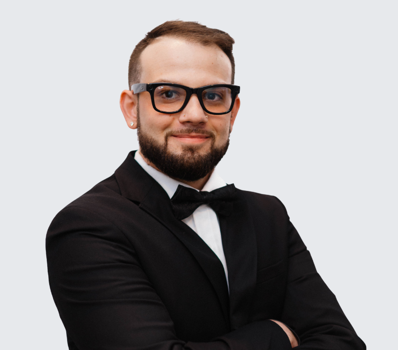 Jake-26 A man with a beard and glasses wearing a black suit and bow tie, with arms crossed.