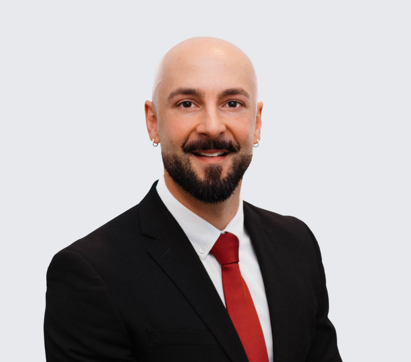 Josh-26 A bald man with a beard and small hoop earrings, wearing a black suit, white shirt, and red tie.