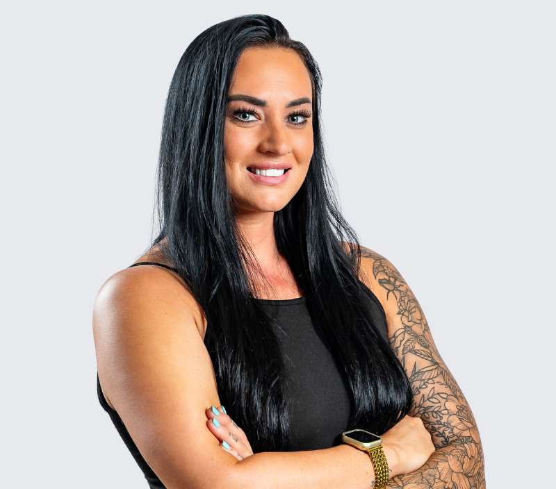 Micheh_Waters A woman with long black hair and a tattooed arm smiling with her arms crossed.