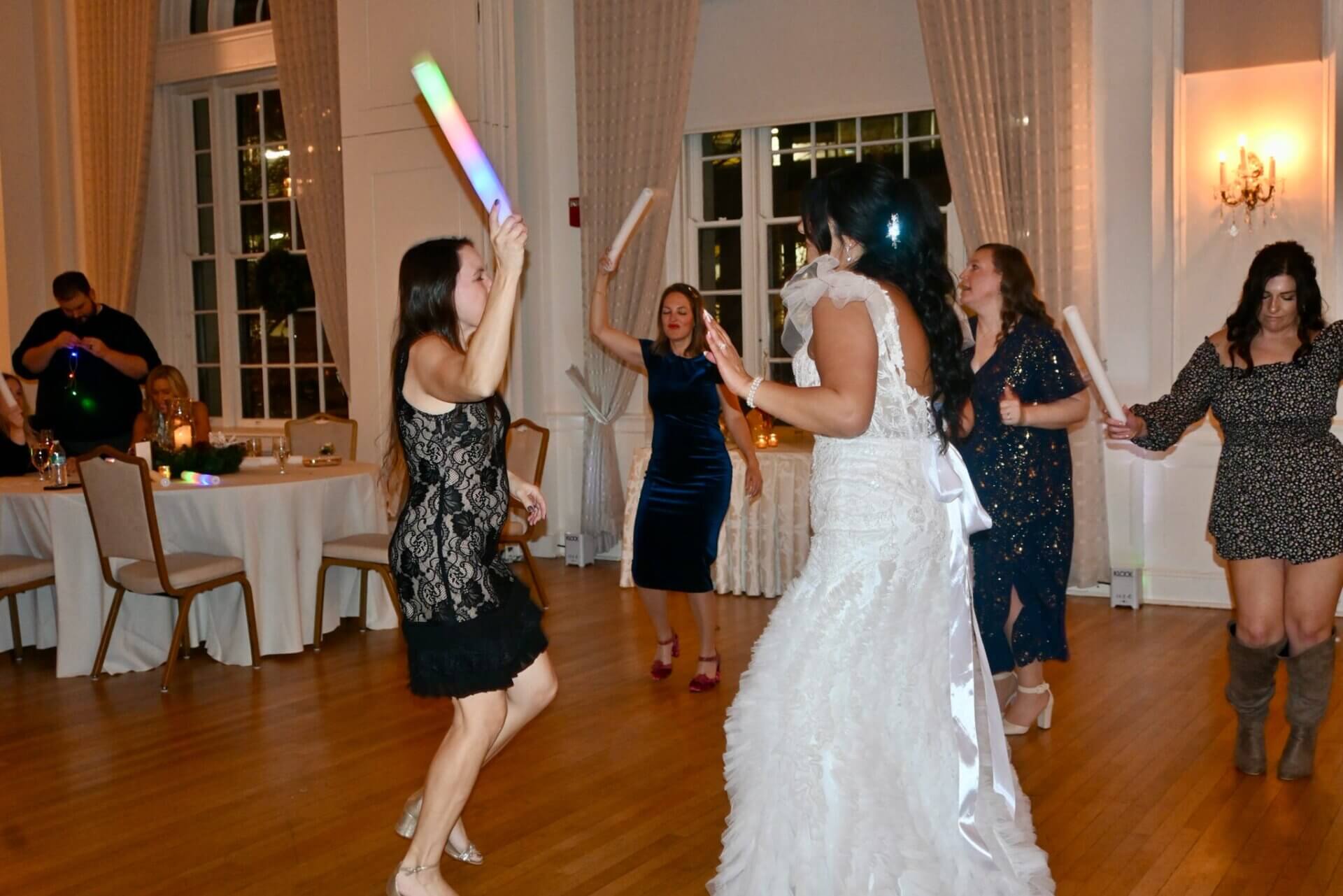 A bride and wedding guests dancing on a wooden floor, holding glowing foam sticks.